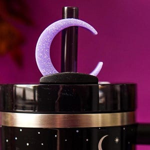 May include: A black tumbler lid featuring a black straw topped with a purple glitter crescent moon accessory. The tumbler has a black exterior decorated with white stars and moon motifs, accented by a metallic silver band near the top rim.
