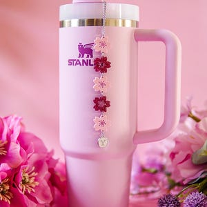 May include: A light pink insulated tumbler with a handle, a white straw, and a silver-toned metal chain charm featuring five alternating light and dark pink flower-shaped acrylic charms and a small circular pendant with the letter Y engraved on it.
