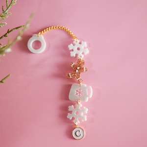 Puede incluir: Un brazalete blanco y dorado con charms de copo de nieve, hombre de jengibre, taza de caf&eacute; y copo de nieve. El brazalete tiene una cadena dorada y un charm circular blanco con la letra "C" en &eacute;l.