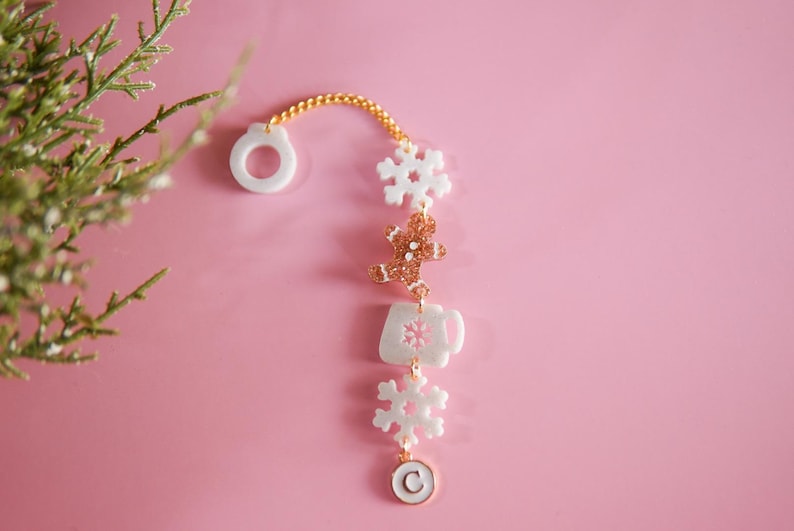 Puede incluir: Un brazalete blanco y dorado con charms de copo de nieve, hombre de jengibre, taza de caf&eacute; y copo de nieve. El brazalete tiene una cadena dorada y un charm circular blanco con la letra "C" en &eacute;l.