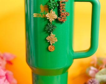 May include: A green tumbler with a handle, a straw, and a brown textured base. A gold chain charm hangs from the lid, featuring glittery green and gold shamrock shapes, a glittery orange script word that reads Lucky, and a round charm with the letter K.