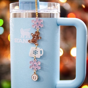 Puede incluir: Un vaso de acero inoxidable azul claro con una cadena dorada y abalorios. Los abalorios incluyen un copo de nieve, un hombre de jengibre, una taza blanca con un copo de nieve, otro copo de nieve y un abalorio dorado con la letra Q. El vaso est&aacute; delante de un fondo borroso de luces navide&ntilde;as.