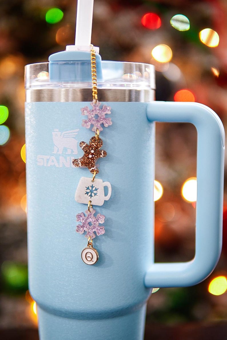 Puede incluir: Un vaso de acero inoxidable azul claro con una cadena dorada y abalorios. Los abalorios incluyen un copo de nieve, un hombre de jengibre, una taza blanca con un copo de nieve, otro copo de nieve y un abalorio dorado con la letra Q. El vaso est&aacute; delante de un fondo borroso de luces navide&ntilde;as.