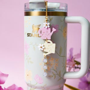 May include: A floral-patterned Stanley mug with a white handle and gold accents. A charm with pink and cream watering can pendants and daisy accents hangs from the lid. The mug is light blue with pink and yellow floral designs.