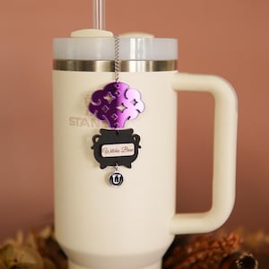 May include: A white travel mug with a purple and black charm hanging from the lid. The charm features a cauldron with a purple smoke cloud and the text "Witches Brew".