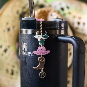 May include: A black Stanley tumbler with a decorative charm. The charm features a cow head, a pink glitter cowboy hat, a brown cowboy boot, and a gold-coloured charm with the letter "W". A cowboy hat is in the background.