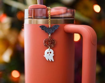 May include: A matte orange tumbler with a handle and a straw, featuring a decorative charm attached to the lid. The charm consists of a gold-tone chain with a black glitter bat, an orange glitter pumpkin, and a white ghost pendant hanging vertically.