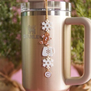 Puede incluir: Un vaso de acero inoxidable dorado con una tapa blanca y una cadena dorada con dijes blancos. Los dijes incluyen un copo de nieve, un hombre de jengibre, una taza de caf&eacute; y un copo de nieve. El vaso tiene la palabra "STANLEY" impresa en el lateral.