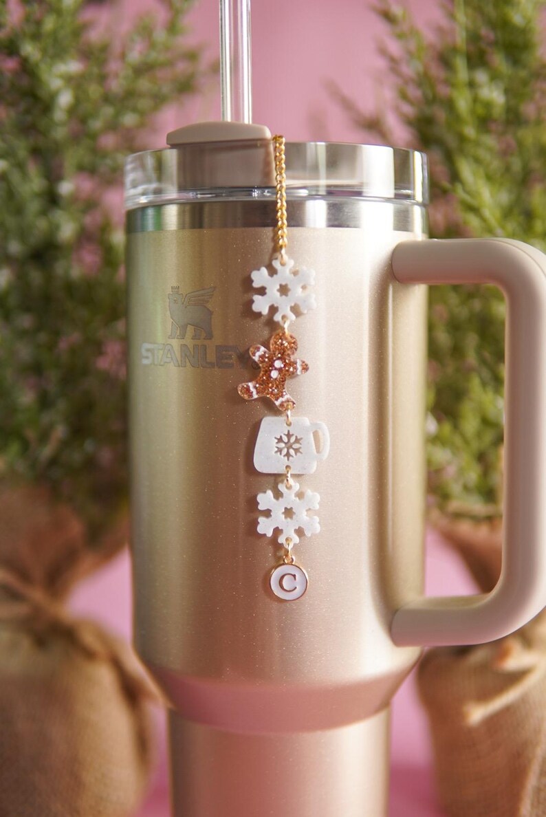 Puede incluir: Un vaso de acero inoxidable dorado con una tapa blanca y una cadena dorada con dijes blancos. Los dijes incluyen un copo de nieve, un hombre de jengibre, una taza de caf&eacute; y un copo de nieve. El vaso tiene la palabra "STANLEY" impresa en el lateral.