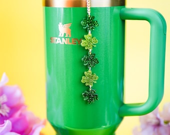 May include: A green Stanley tumbler with a handle and a straw, featuring a vertical charm chain attached to the lid. The chain consists of five glittery, four-leaf clover shapes in varying shades of green, hanging down the front of the cup.