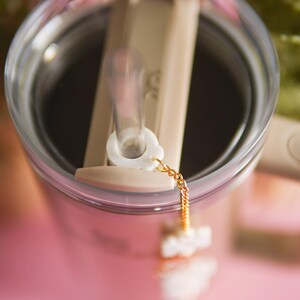 Puede incluir: Primer plano de un vaso de vidrio transparente con tapa beige y una pajita. La pajita tiene un tap&oacute;n blanco y una cadena dorada unida a ella. El vaso est&aacute; sobre una superficie rosa.