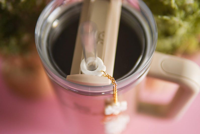 Puede incluir: Primer plano de un vaso de vidrio transparente con tapa beige y una pajita. La pajita tiene un tap&oacute;n blanco y una cadena dorada unida a ella. El vaso est&aacute; sobre una superficie rosa.