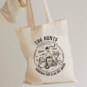 May include: A cream-coloured tote bag featuring black graphic text and illustrations. The text reads "The Aunts Midnight Margaritas since 1998" with images of a cauldron, tequila bottle, margarita glass, and a pumpkin.
