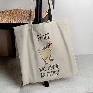 Könnte beinhalten: Beige Canvas-Tragetasche mit einer Cartoon-Gans, die ein Messer hält. Der Text "PEACE WAS NEVER AN OPTION" ist in Schwarz über und unter der Gänse-Illustration gedruckt. Die Tasche hat zwei lange Henkel.