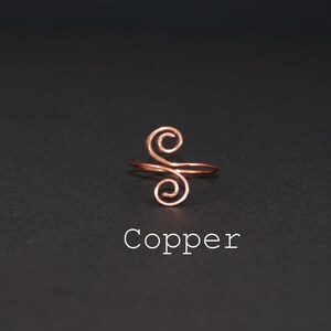 Op de afbeelding: Een koperen draadring met een dubbel swirl-ontwerp. De ring ligt op een zwarte achtergrond en het woord "Koper" staat onder de ring.