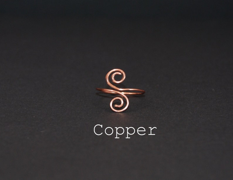 Op de afbeelding: Een koperen draadring met een dubbel swirl-ontwerp. De ring ligt op een zwarte achtergrond en het woord "Koper" staat onder de ring.