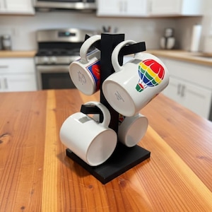 May include: A black wooden mug tree holds four white ceramic mugs. One mug features a colorful hot air balloon design. The other mugs are plain white. The mug tree is on a wooden surface.