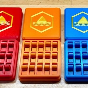 May include: Three colorful, 3D-printed storage boxes. Each box has a lid with a gold hexagon design featuring a shark fin and waves. The boxes are red, orange, and blue, with matching colored interiors designed to hold small items.