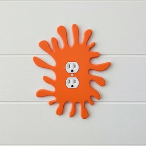 May include: Orange splash-shaped outlet cover with two white electrical outlets. The cover is mounted on a white, horizontally-paneled wall. The splash design adds a playful touch to a functional item.