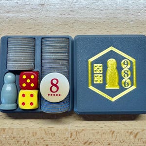 May include: A gray board game organizer with compartments holding game pieces. Includes stacks of gray tokens, red and yellow dice, a white token with the number 8, and a gray game piece. The lid features a gold emblem with dice and numbers.