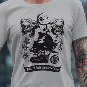 May include: A gray t-shirt with a black and white graphic design of a ship sailing on a stormy sea. Two skulls with bandanas are on either side of the ship. The text "Ship of fools on a cruel sea" is printed below the graphic.