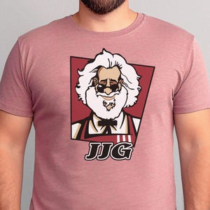 May include: A dusty rose t-shirt featuring a cartoon illustration of a man with white hair, sunglasses, and a white beard, in a red and black frame. The text "JJG" is below the image. The shirt is a solid color.