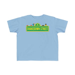 May include: A light blue t-shirt featuring a green and yellow street sign design with the words "SHAKEDOWN STREET" in white. The design includes cartoon characters and a yellow heart. The shirt is made of a soft material.