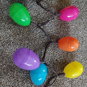 May include: A string of Easter egg-shaped lights in various colors, including blue, green, yellow, pink, orange, and purple. The lights are connected by a brown wire, creating a festive and colorful display.