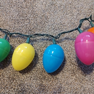 May include: A string of colorful plastic Easter egg lights. The lights are in the shape of eggs and are strung together with green wire. The eggs are in the colors of orange, green, yellow, blue, and pink.