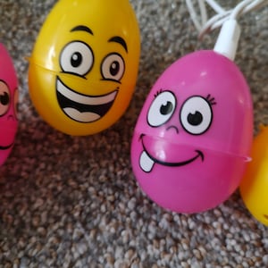 May include: A string of colorful plastic Easter eggs with light bulbs inside. Each egg has a different facial expression. The eggs are yellow, pink, and blue.