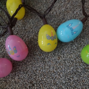 May include: A string of Easter egg lights with unicorn designs. The eggs are in pink, yellow, blue, and green. The lights are strung on brown wire.