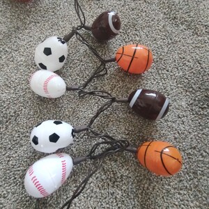 May include: A string of lights with sports-themed light bulbs. The bulbs are shaped like baseballs, basketballs, footballs, and soccer balls. The lights are strung together with brown wire.