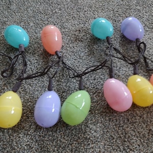 May include: A string of pastel colored plastic Easter egg lights. The lights are connected by a black wire and have a variety of colors including light blue, pink, yellow, green, and purple.