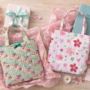 Könnte beinhalten: Zwei Tragetaschen mit Blumenmuster, eine mit einem bunten Muster und die andere mit rosa Blumen und Herzen, liegen auf rosa Seidenpapier. Eine Geschenkbox mit einer türkisfarbenen Schleife, eine Bibel und ein Strauß rosa Rosen sind ebenfalls vorhanden.