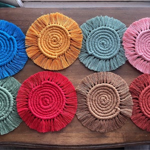 May include: Eight colorful crocheted coasters with fringe. The coasters are in shades of blue, orange, green, pink, red, and brown.