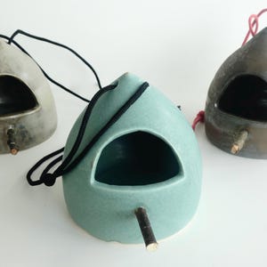 May include: Three ceramic birdhouses in grey, teal, and brown. Each has a rounded shape with a circular opening and a small wooden perch. Black or red cords are attached for hanging.