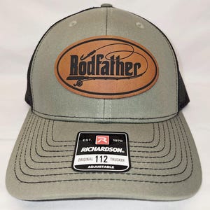 Może przedstawiać: Szara i czarna czapka trucker z brązową skórzaną naszywką z napisem "Rodfather" i wzorem wędki i żyłki. Czapka jest regulowana i ma logo Richardson z tyłu.