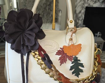 Op de afbeelding: Een crèmekleurige Steve Madden handtas met een grote donkerbruine stoffen bloem, een goudkleurige kettingriem verweven met veelkleurige stoffen stroken en een decoratieve bedelcluster van oranje, bordeauxrode en donkergroene bladvormen.