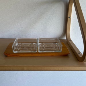 May include: Two clear glass dishes with a decorative pattern sit on a wooden tray. The tray is on a light wood shelf.