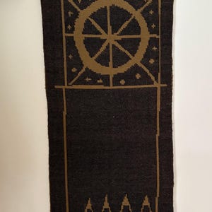 Puede incluir: Un tapiz de pared tejido de color marrón oscuro con un diseño geométrico en color beige. El diseño incluye una rueda, arcos y círculos. El tapiz de pared es rectangular y cuelga de una barra de metal negro.