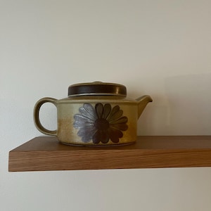 May include: A ceramic teapot with a brown lid and handle, featuring a large floral design in shades of brown and green. The teapot is set on a wooden shelf against a white wall. A vintage kitchenware item.