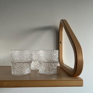 Könnte beinhalten: Drei klare Glasbecher mit strukturierter Oberfläche stehen auf einem Holzregal. Das Regal ist mit einer Holzhalterung an einer Wand befestigt.
