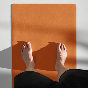 Puede incluir: Una persona de pie sobre una gran estera de yoga naranja con los pies descalzos. La estera está sobre una superficie blanca.