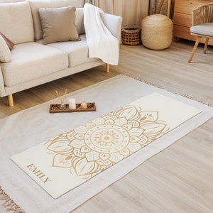 Puede incluir: Una esterilla de yoga blanca con un diseño de mandala dorado y el nombre "EMILY" impreso en la esquina inferior izquierda.