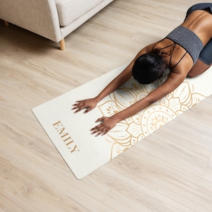 Puede incluir: Una esterilla de yoga blanca con un diseño de mandala dorado y el nombre "EMILY" impreso en ella. Una persona está haciendo yoga en la esterilla.