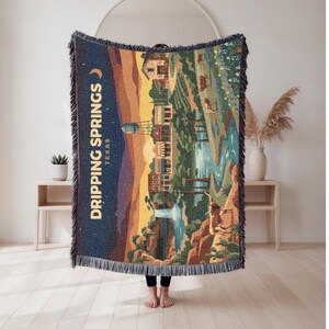 May include: A woven throw blanket featuring a landscape illustration of Dripping Springs, Texas. The design includes a dark blue night sky with a crescent moon, a water tower, buildings, a river, and cattle. The blanket has fringed edges.