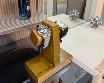 May include: A wooden watch stand holding two wristwatches. One watch has a silver strap and a black face, the other has a black leather strap and a white face. A bottle of cologne is on a shelf. The text "YAKUP KAÇAR" is visible on the stand.