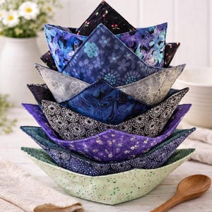 May include: A stack of handmade fabric bowl cozies in various shades of blue, purple, and green, each with a unique floral pattern. The cozies are arranged in a nested fashion, showcasing their triangular shape. A wooden spoon and a white vase with flowers are in the background.