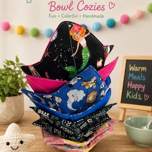 May include: A stack of five fabric bowl cozies featuring various patterns including a mermaid on a black background, a blue animal print with elephants, a black and white text print, and a pink and yellow floral print with quilted stitching.