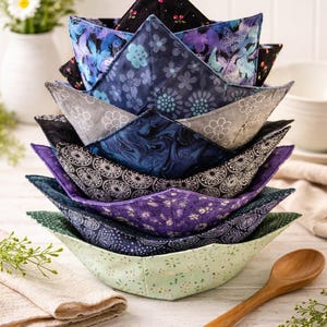 May include: A stack of eight fabric bowl cozies featuring various patterns including black with cherries, blue floral, grey with white flowers, dark blue marble, black and white geometric, purple floral, navy polka dot, and light green floral.